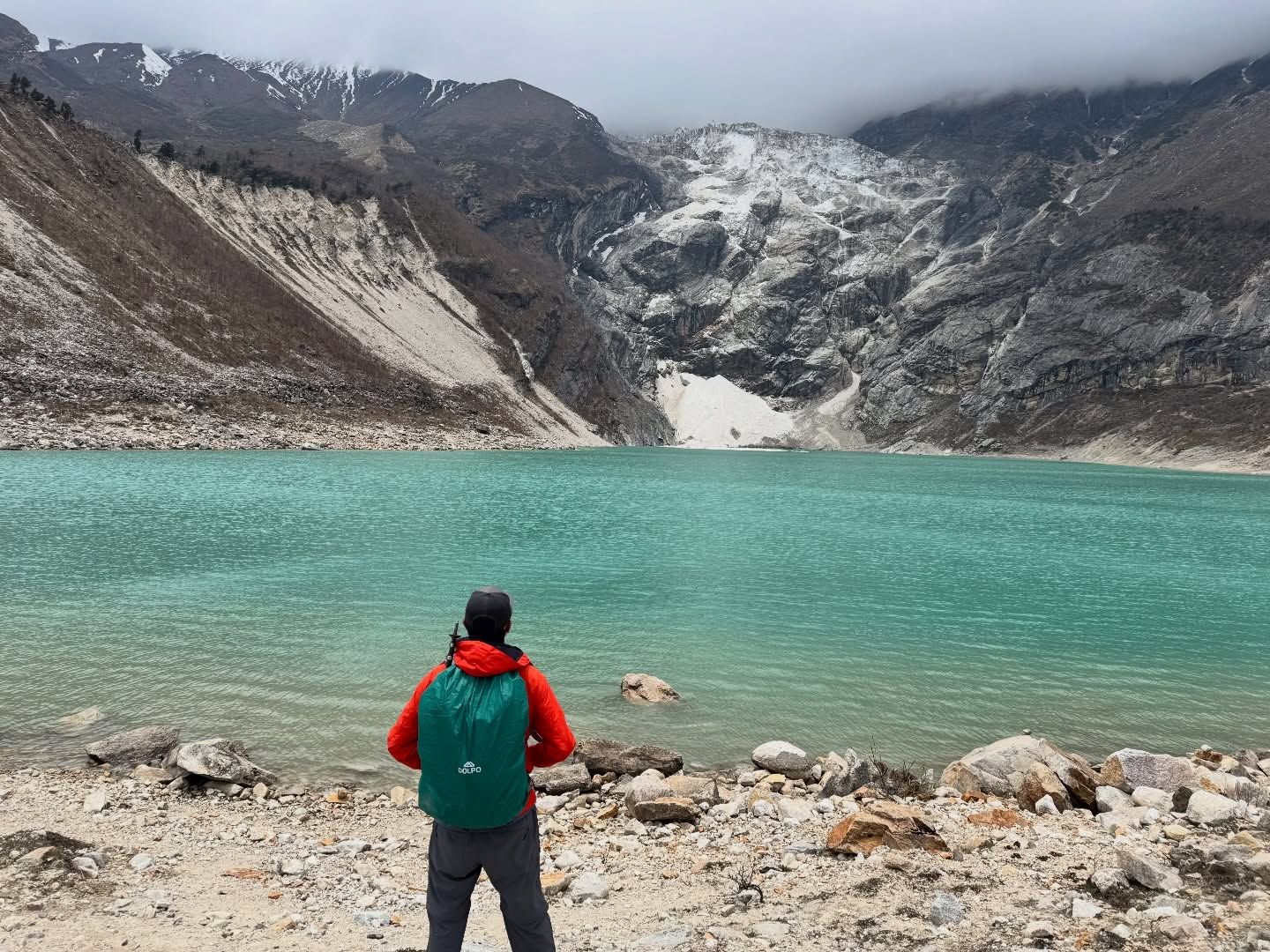 Oshan from behind, in a red jacket with a green pack, looking across the turquoise water of Birendra Tal to a glacier under low cloud.