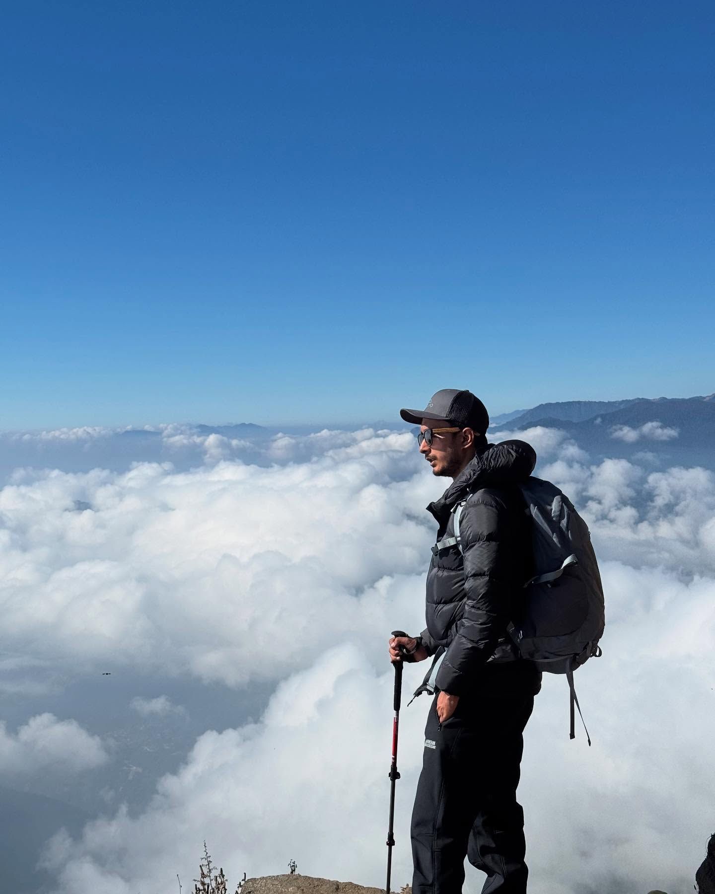 Oshan in a black down jacket with a trekking pole, standing above a sea of white cloud.