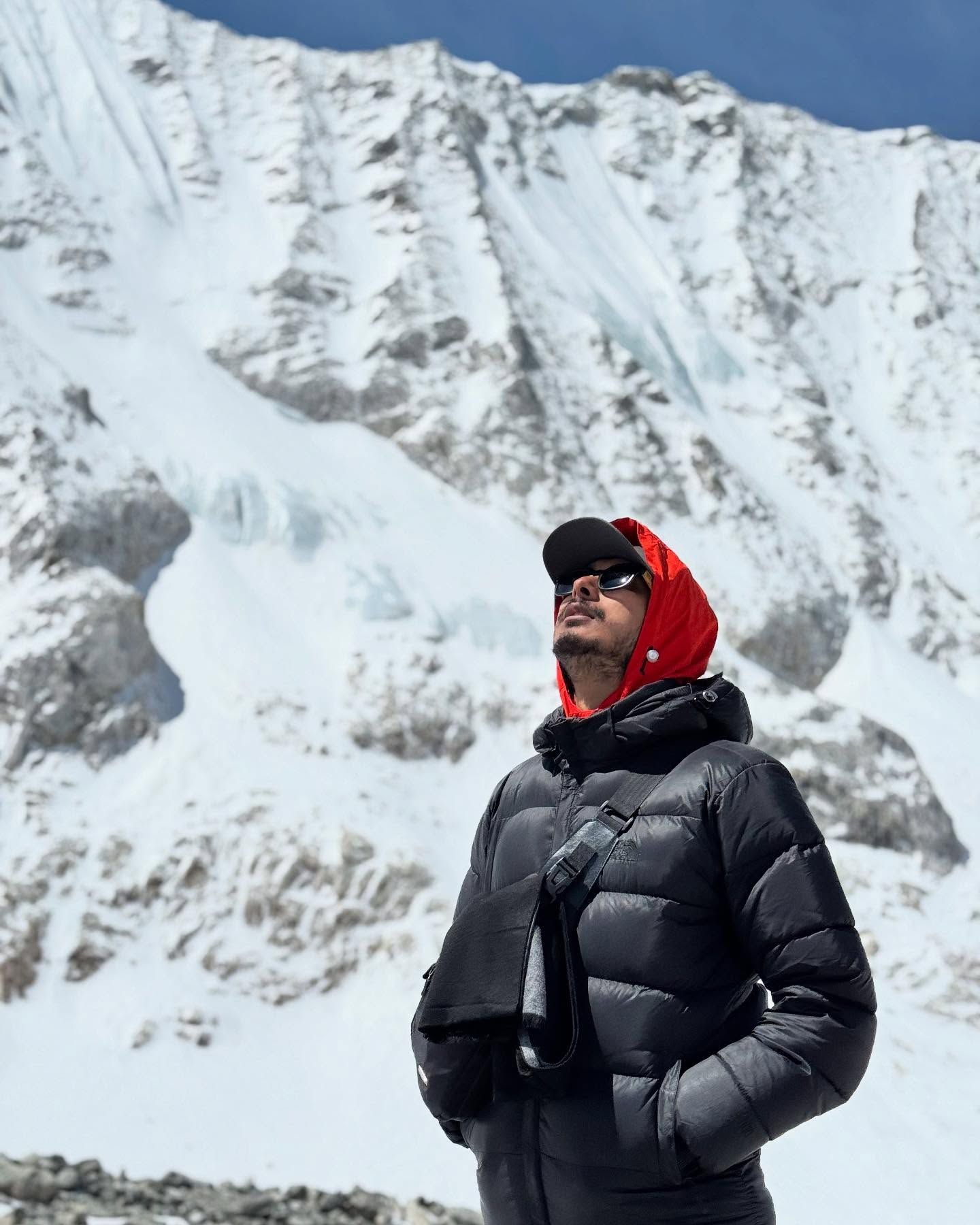 Oshan in a black down jacket, red hood and sunglasses, looking up in front of a steep face of snow and ice.
