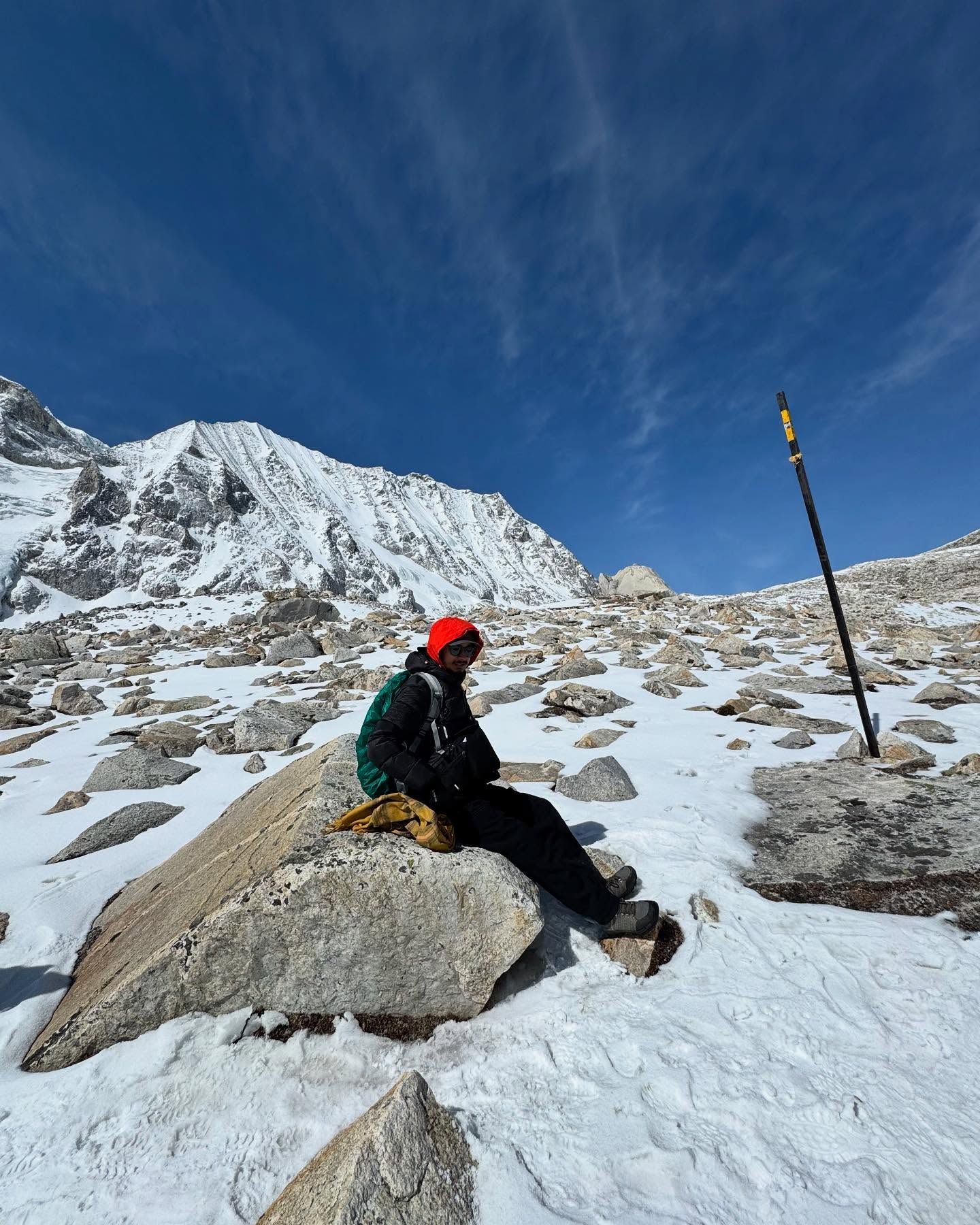 Oshan resting on a boulder in the snow beside a black trail-marker pole, with a snow-covered peak behind.