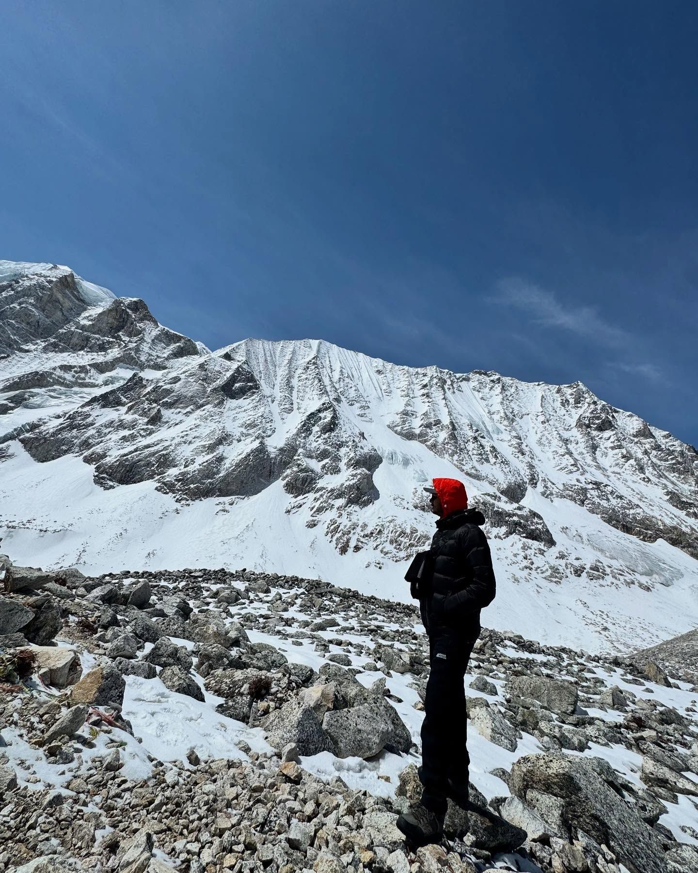 Oshan in a black down jacket and red hood, standing on snowy boulders below a long snow-covered ridge under a deep blue sky.