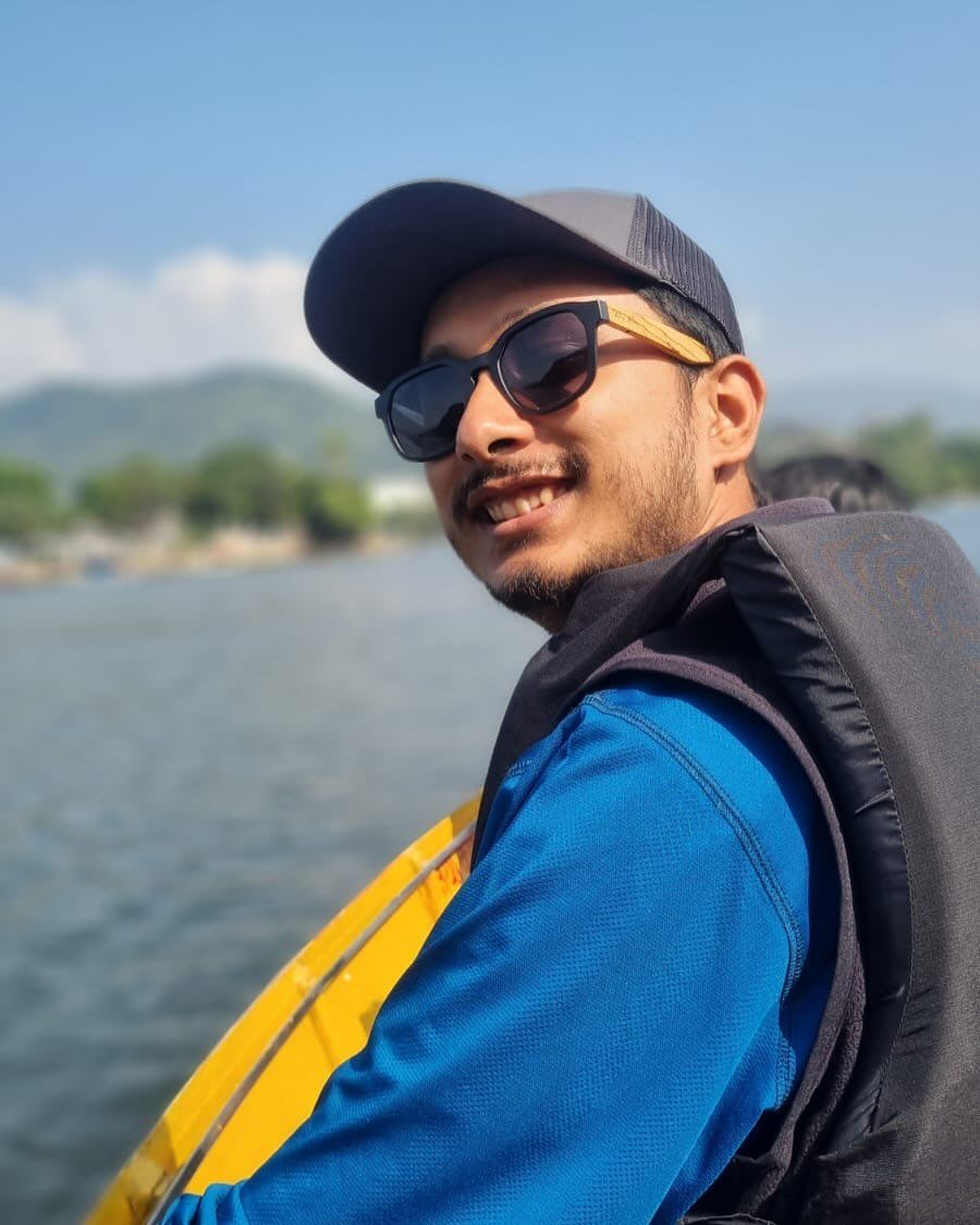 Oshan in a cap, sunglasses and a life jacket, smiling back over a shoulder from a yellow boat on Phewa Lake.