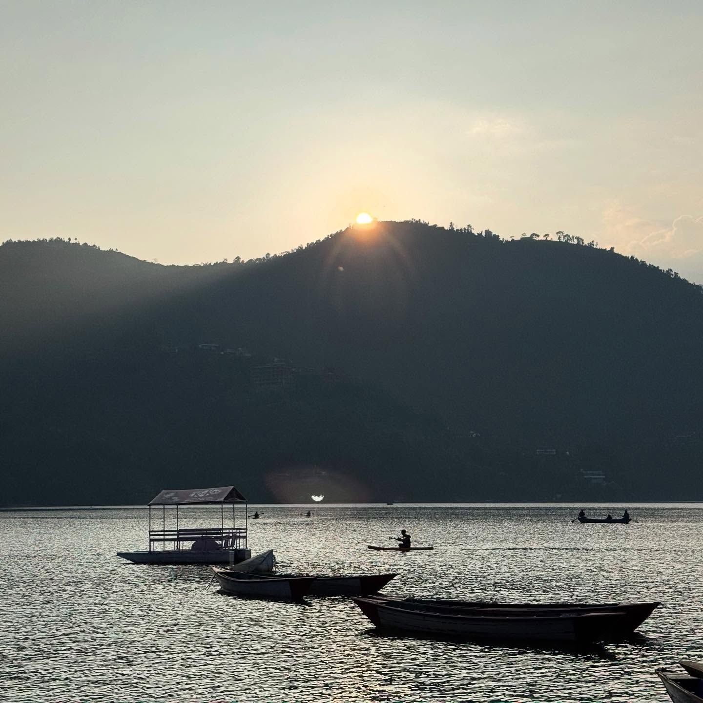 The sun setting over a dark hill above Phewa Lake, with boats moored on the water.