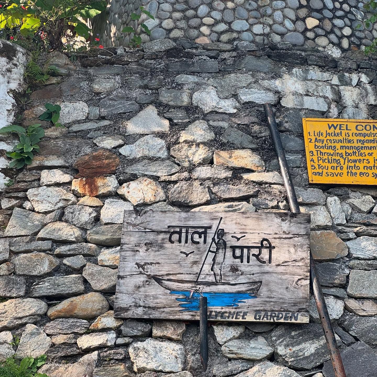 A weathered wooden sign reading "ताल पारी" (across the lake), with a painted boat, set in a stone wall.