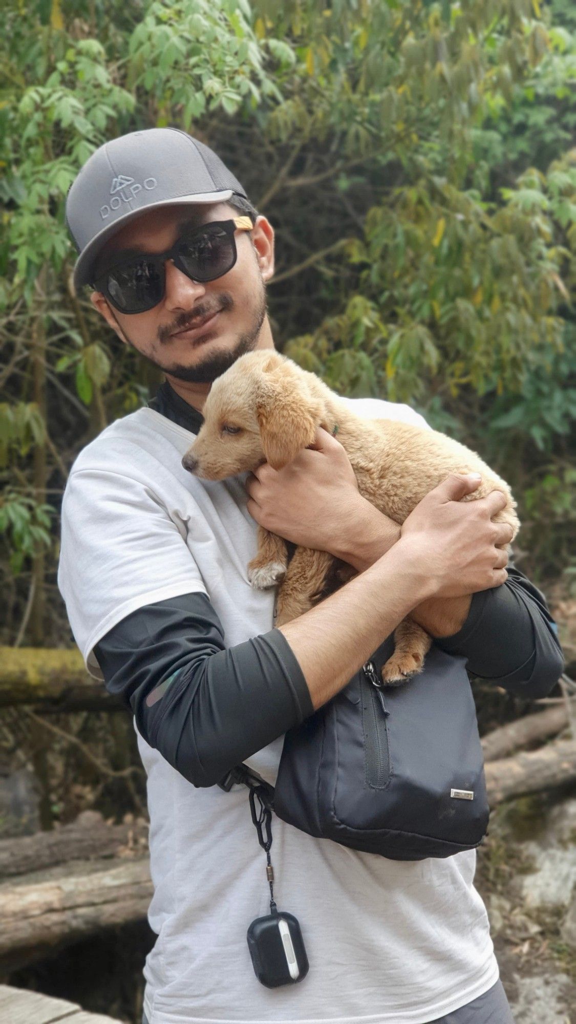 Oshan in a grey cap and sunglasses, smiling and holding a sleepy golden puppy, with green forest behind.