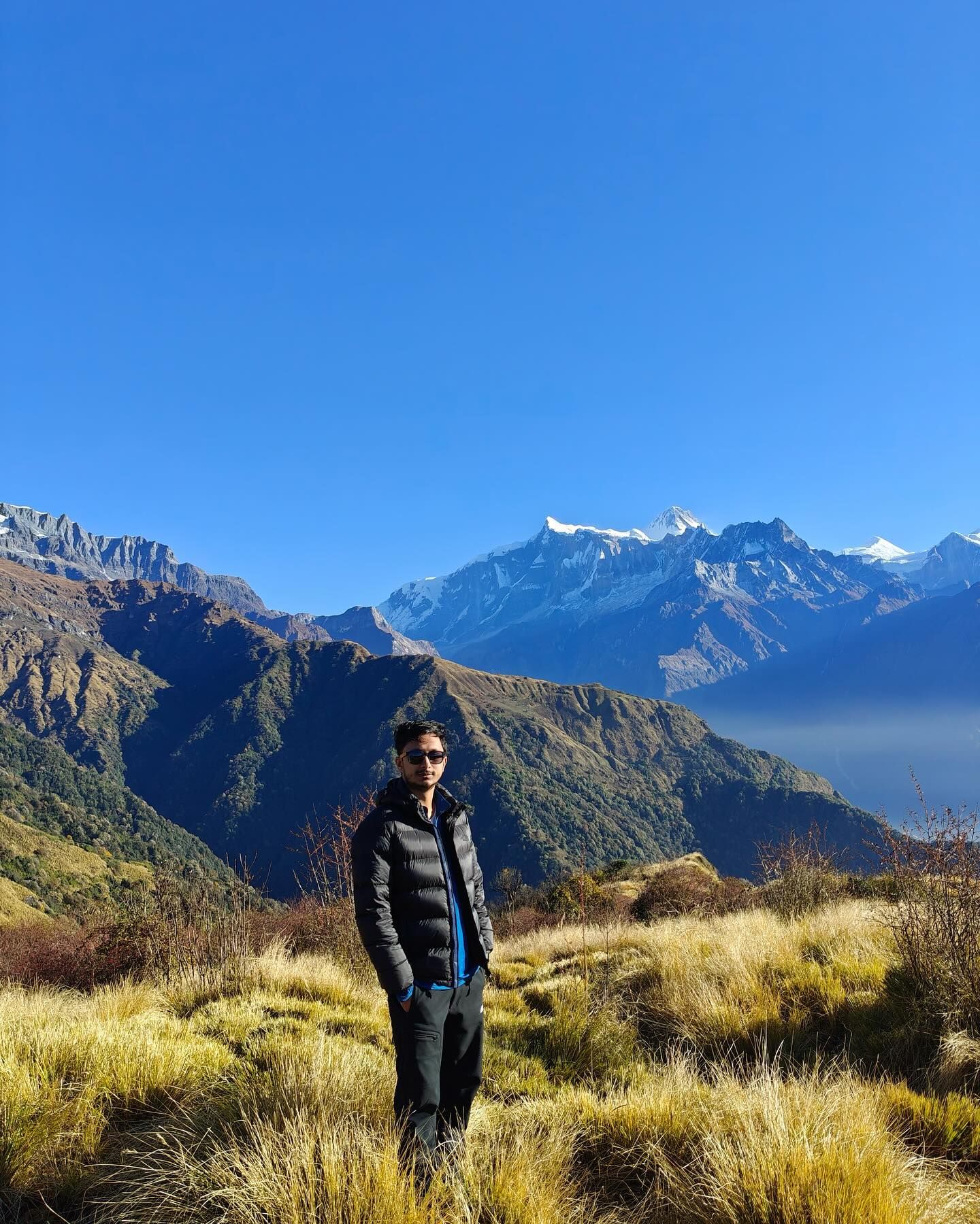 Oshan in a black down jacket on a slope of golden grass, with forested ridges and snow peaks behind.